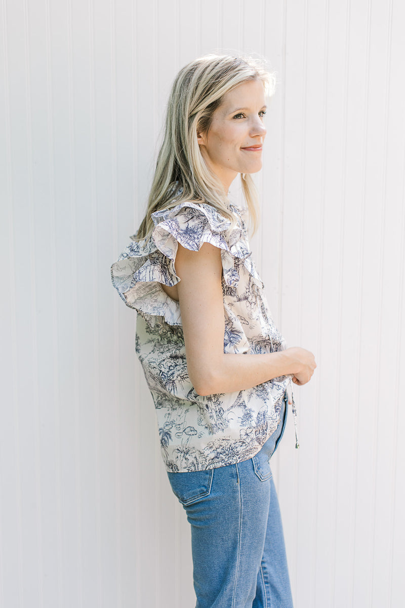 Side view of Model wearing a blue toile top with layered ruffle cap sleeves and a v-neck.