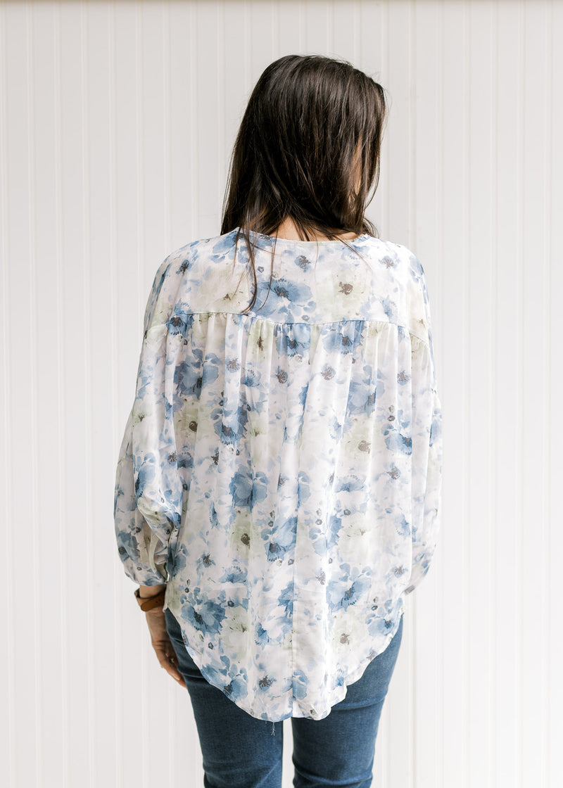 Back view of Model wearing a white top with a blue floral print, sheer long sleeves and tie at neck.