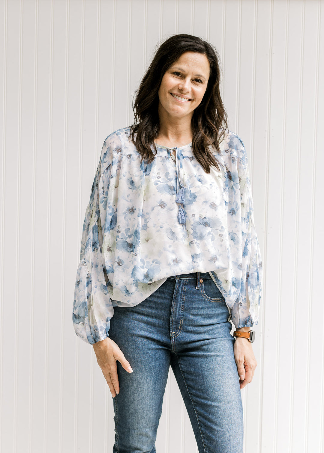 Model wearing a white top with a blue floral watercolor print, sheer long sleeves and tie at neck.