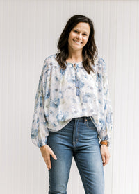 Model wearing a white top with a blue floral watercolor print, sheer long sleeves and tie at neck.