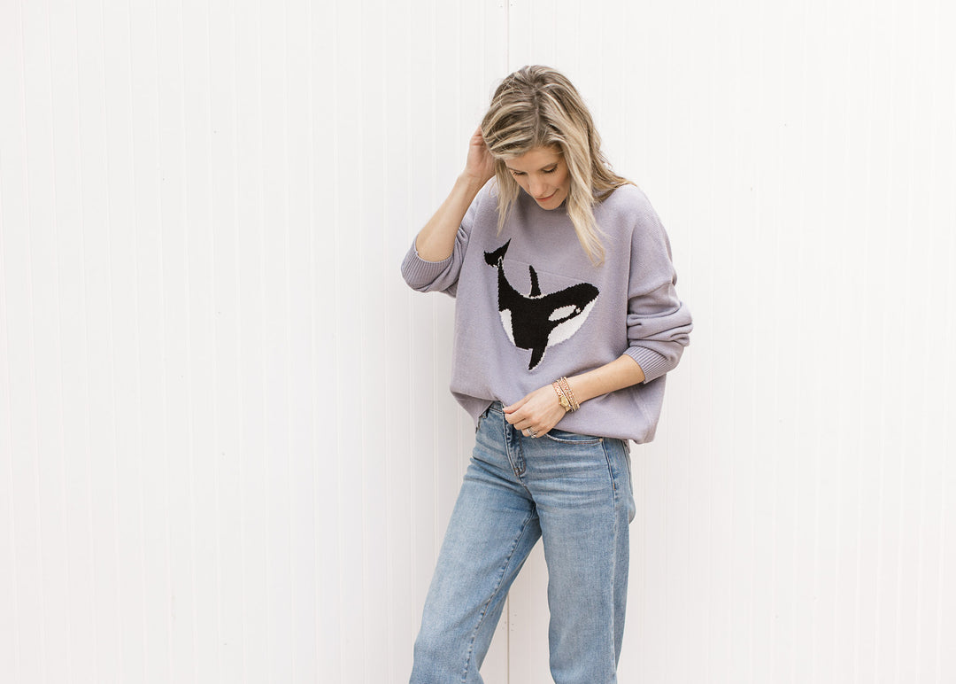 Model wearing jeans and a purplish-blue sweater with a whale graphic, round neck and long sleeves. 