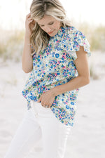 Model wearing a white top with a blue floral pattern, ruffle mock neck, cap sleeves and keyhole. 