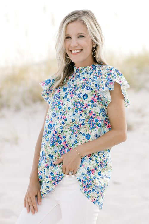 Model wearing a white top with a blue floral pattern, ruffle mock neck and cap sleeves. 