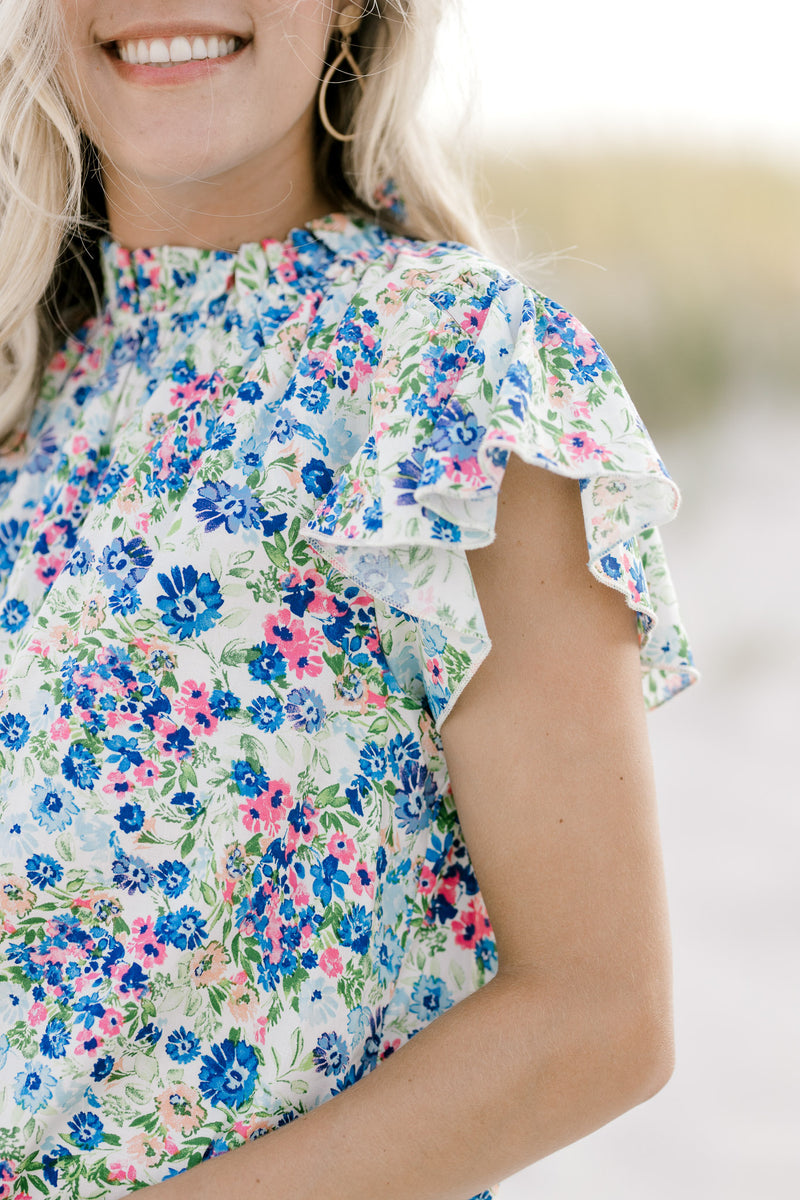Close up view of ruffle mock neck and ruffle cap sleeves on a white top with a blue floral pattern. 
