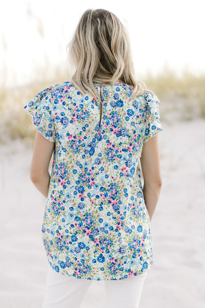 Back view of a model wearing a white top with a blue flora pattern and a keyhole closure. 