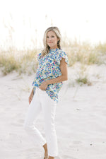 Model wearing white jeans with a white top with a blue floral pattern, mock neck and cap sleeves. 
