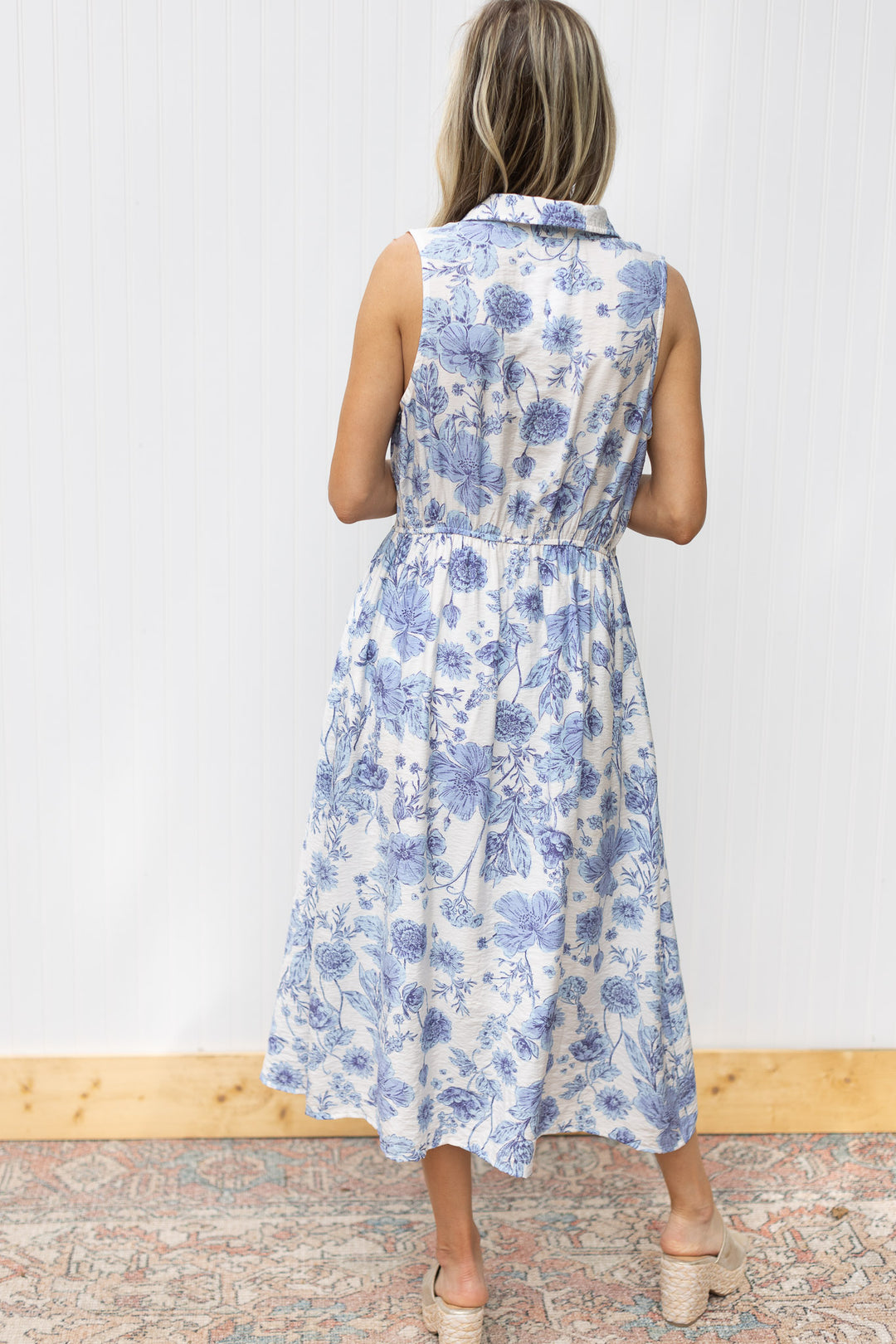 Back view of Model wearing a fully lined white dress with blue floral, button front and a collar.