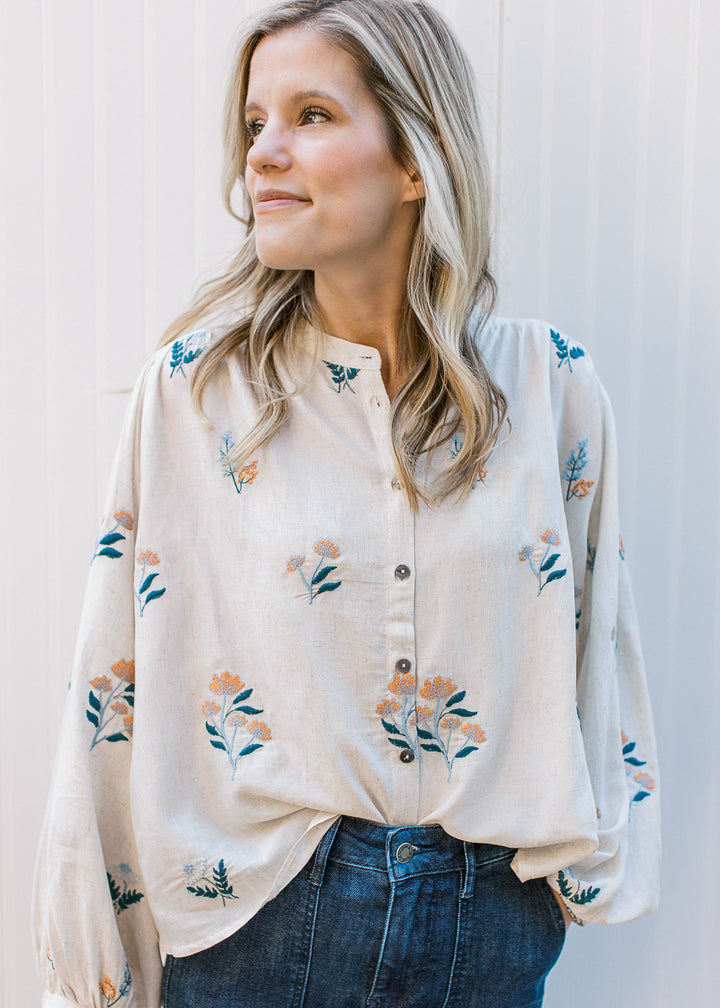 Model wearing an oatmeal top with blue and golden embroidered flowers with bubble long sleeves.