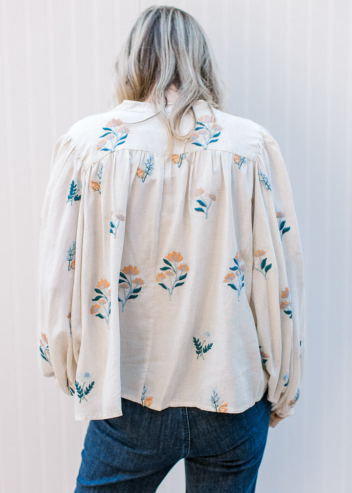 Back view of Model wearing an oatmeal long sleeve top with blue and golden embroidered flowers. 