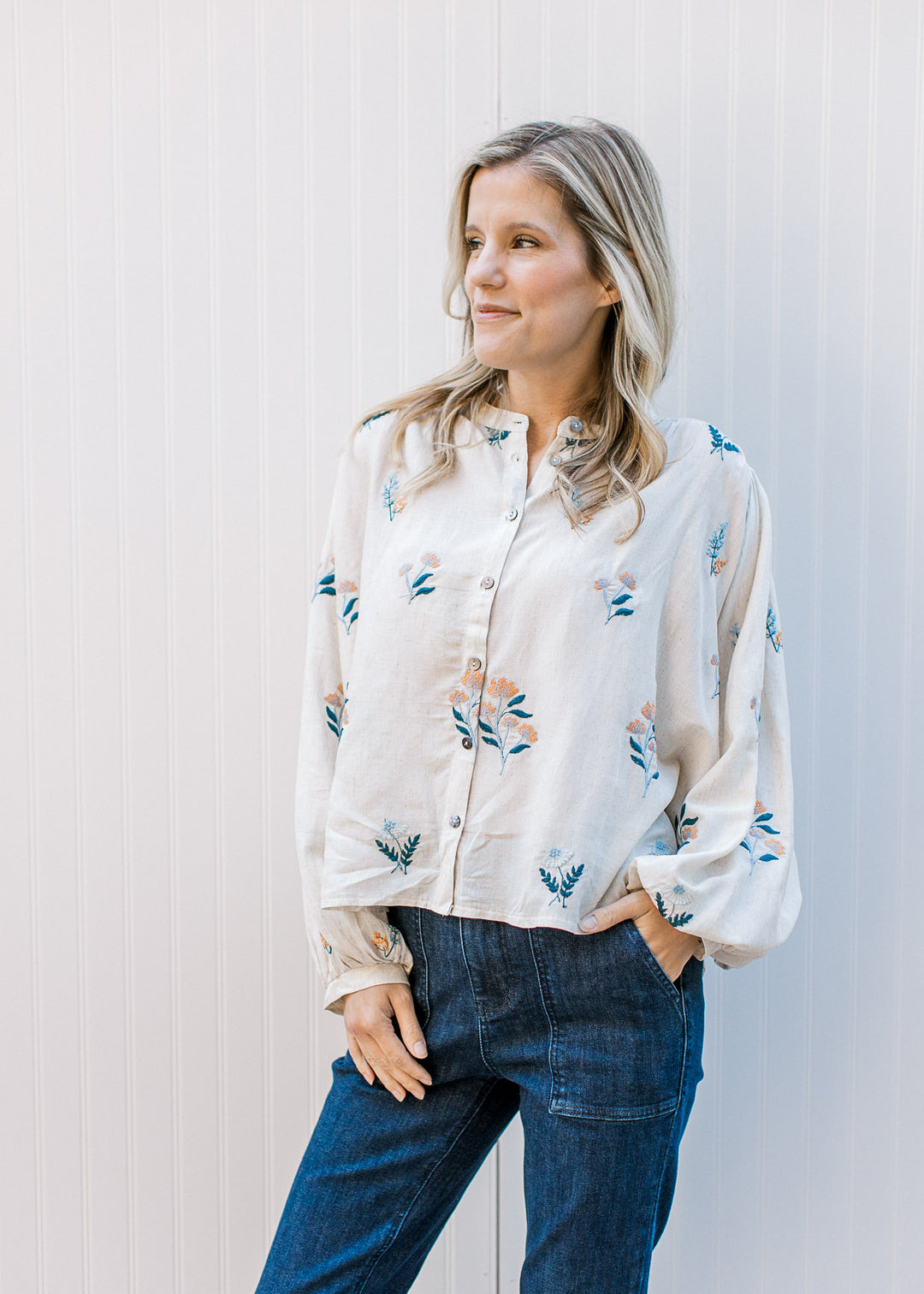 Model wearing jeans and an oatmeal button up with blue and golden embroidered flowers. 
