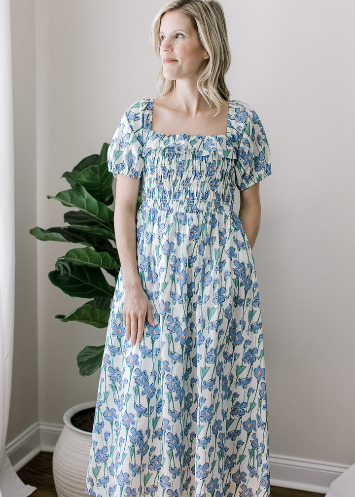 Model wearing a cream midi with blue floral, bubble short sleeves, pockets, and smocked bodice.