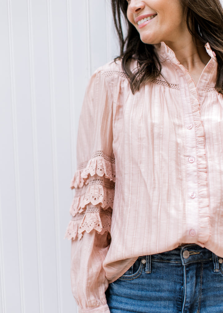Model wearing a blush button up top with mock neck and tiered ruffle detail at elbow of long sleeve.