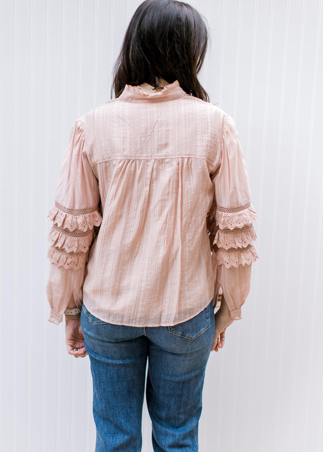 Back view of Model wearing a blush top with mock neck and tiered ruffles at elbow of long sleeve. 