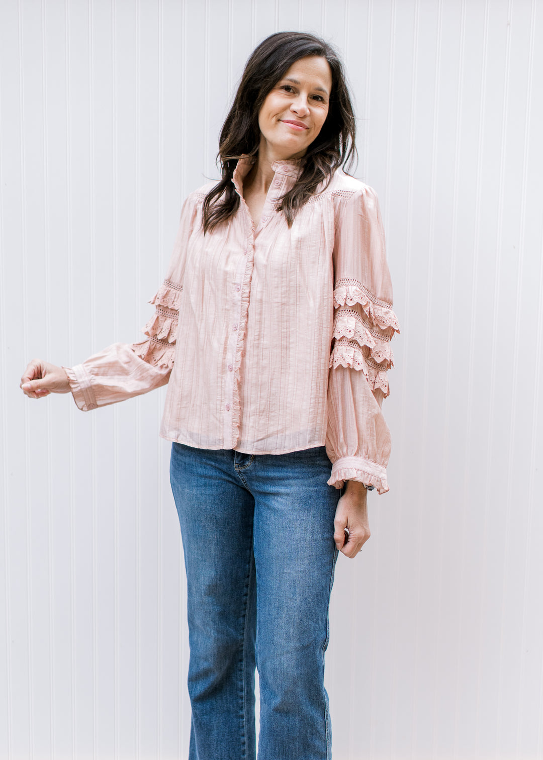 Model wearing jeans and a blush top with mock neck and tiered ruffles at elbow of long sleeve. 