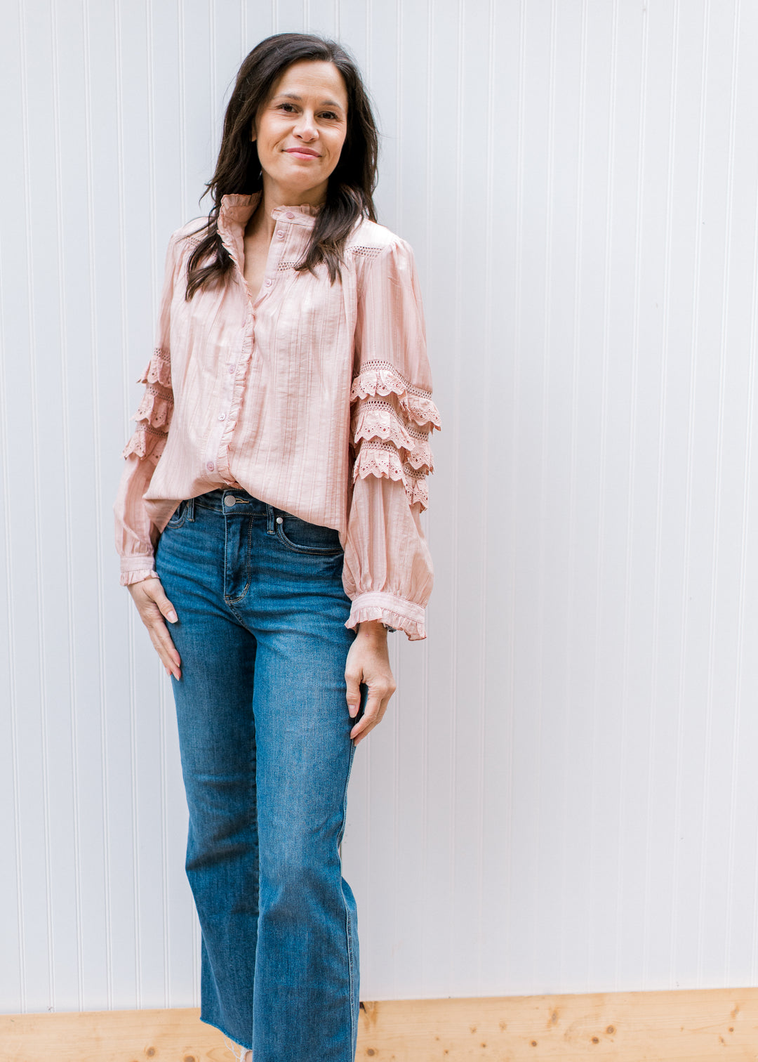 Model wearing a lightweight blush top with mock neck and tiered ruffles at elbow of long sleeve. 