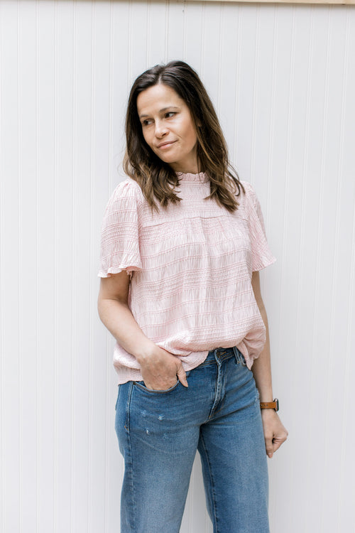 Model wearing a blush top with a ruffled mock neck, short sleeves and a textured material. 