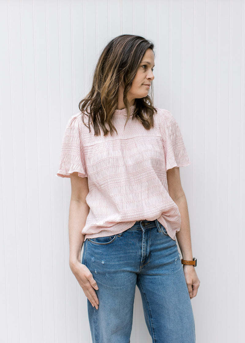 Model wearing jeans and a blush top with a ruffled mock neck, short sleeves and a textured material.