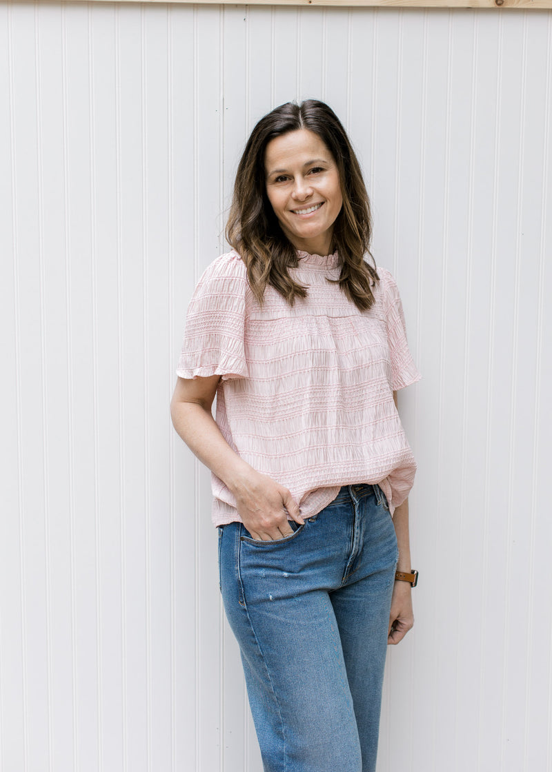 Model wearing a blush top with a ruffled neck, short sleeves and a textured material.