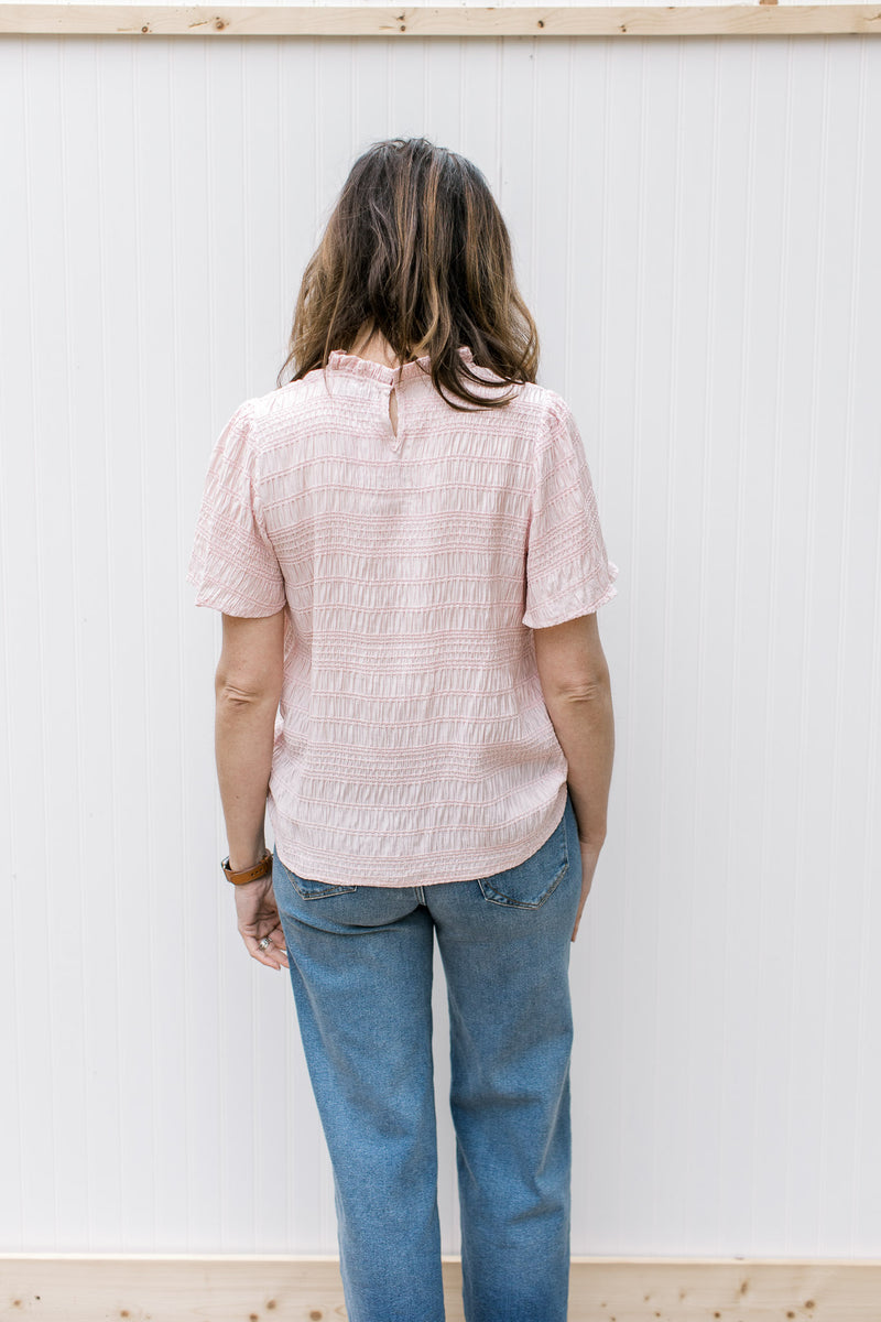 Back view of Model wearing a textured blush top with a ruffled mock neck and short sleeves.