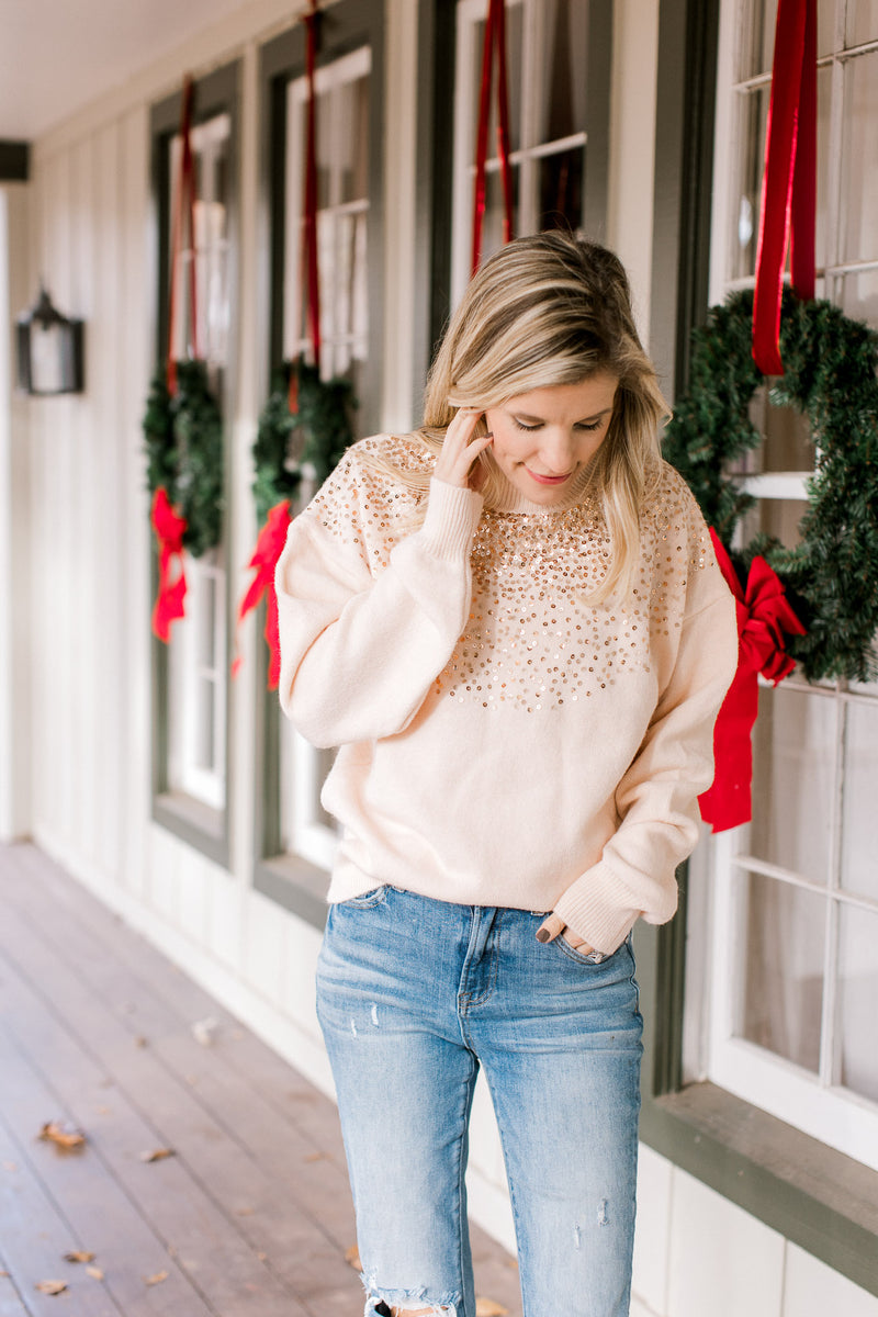 Model wearing jeans with a blush colored sweater with gold sequins at the mock neck and long sleeve.