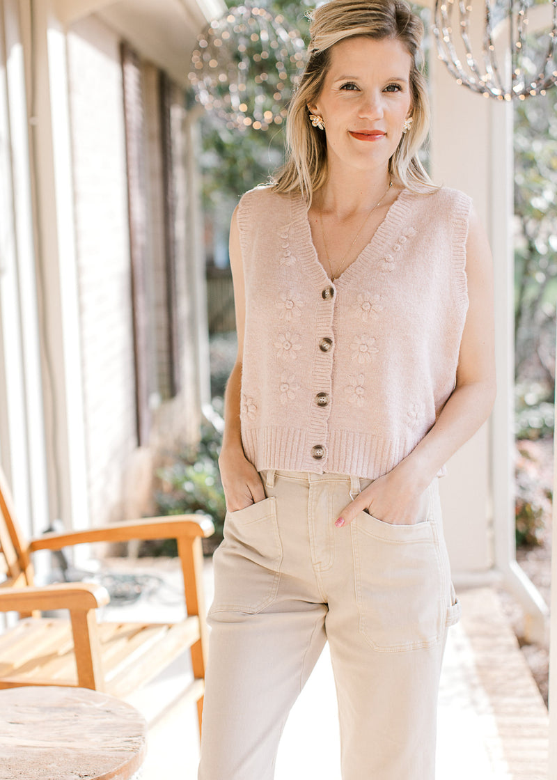 Model wearing khakis and a blush sweater vest with embroidered flowers, v-neck and button front. 