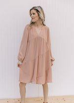 Model wearing a blush v-neck dress with long sleeves and tiers with a cross hem