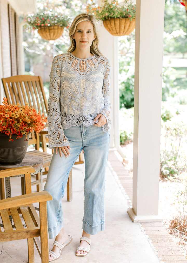 Model wearing jeans and a pale blue and cream open knit sweater with a scallop hem and long sleeves.