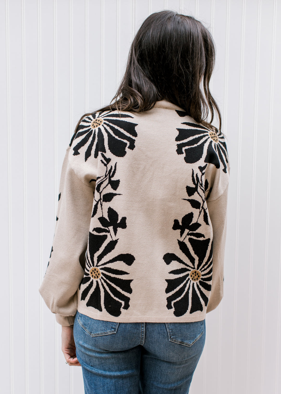 Back view of Model wearing a tan sweater with black floral pattern, a mock neck, and long sleeves. 
