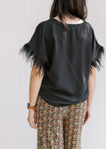 Back view of a Model wearing a black v-neck top with a feather accent on the short sleeves. 