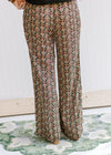 Back view of Model wearing black wide leg pants with a gold, rose and green pattern and pockets. 