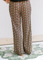 Back view of Model wearing black wide leg pants with a gold, rose and green pattern and pockets. 