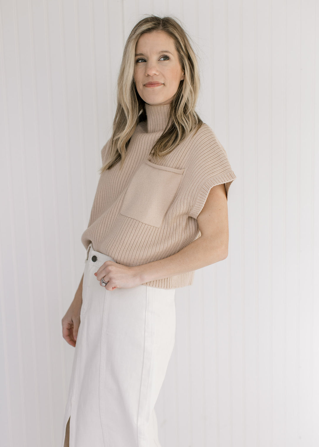 Model wearing a tan cable knit sweater vest with a patch pocket and a mock neckline. 