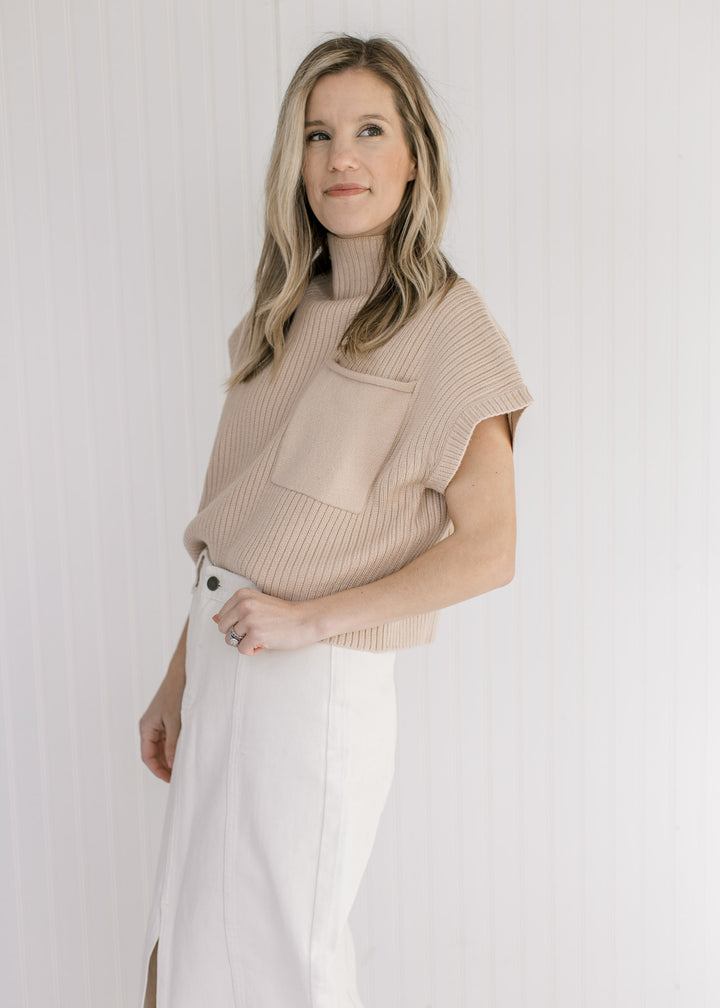 Model wearing a tan cable knit sweater vest with a patch pocket and a mock neckline. 