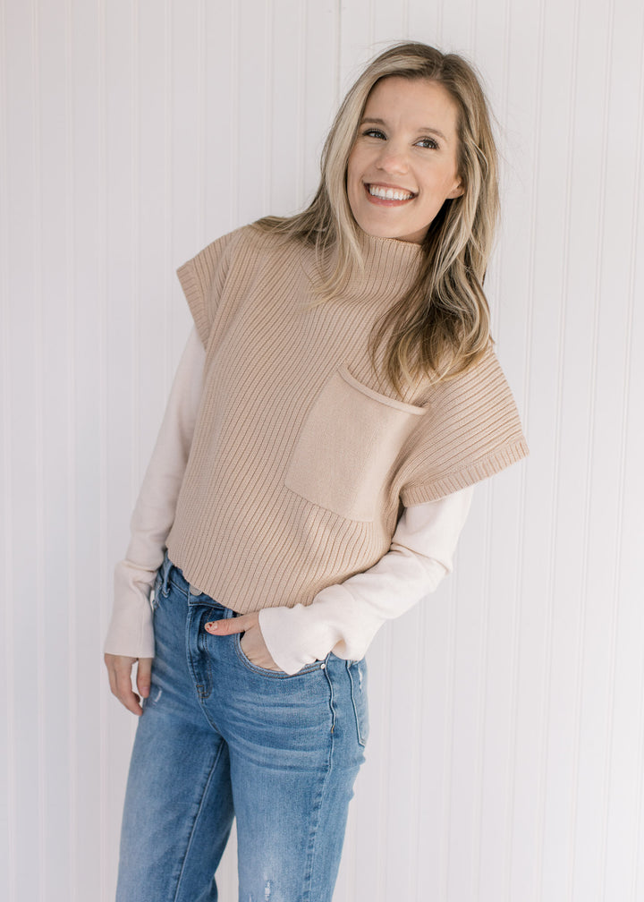 Model wearing a tan cable knit sweater vest over a cream long sleeve top.