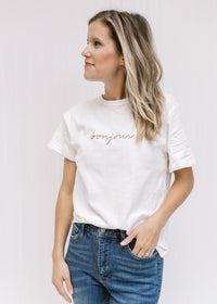 Model wearing a white short sleeve tee with the word bonjour embroidered across the bust in tan. 