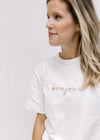 Close up of Model wearing a white tee with the word bonjour embroidered across the bust in tan. 
