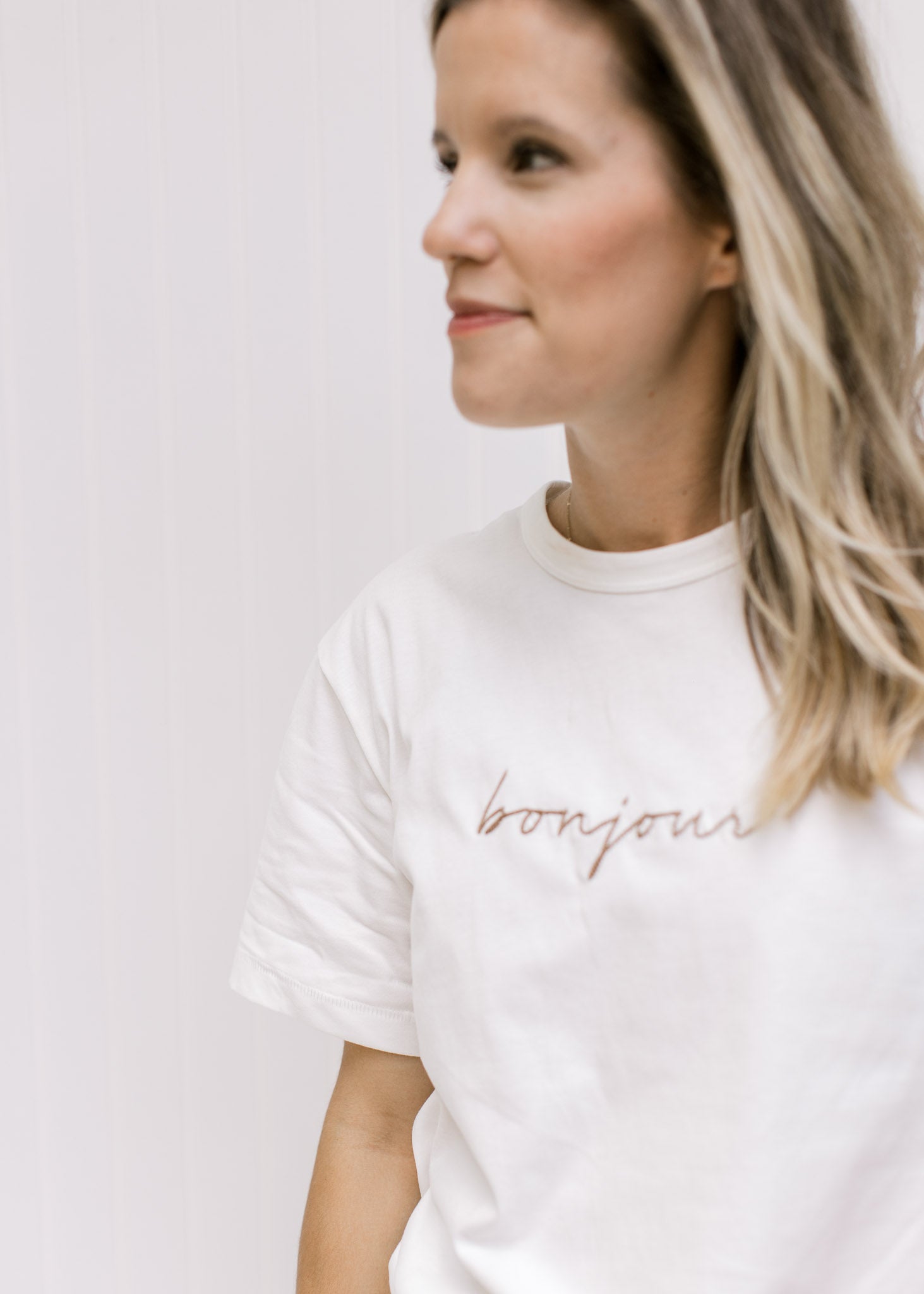 Close up of Model wearing a white tee with the word bonjour embroidered across the bust in tan. 