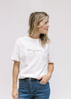 Model wearing jeans with a white short sleeve tee with bonjour embroidered across the bust in tan. 
