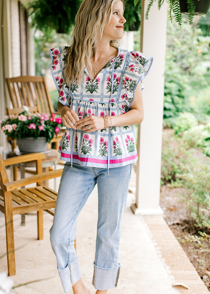 Model wearing an off white top with pale blue squares holding pink flowers and ruffle cap sleeves.