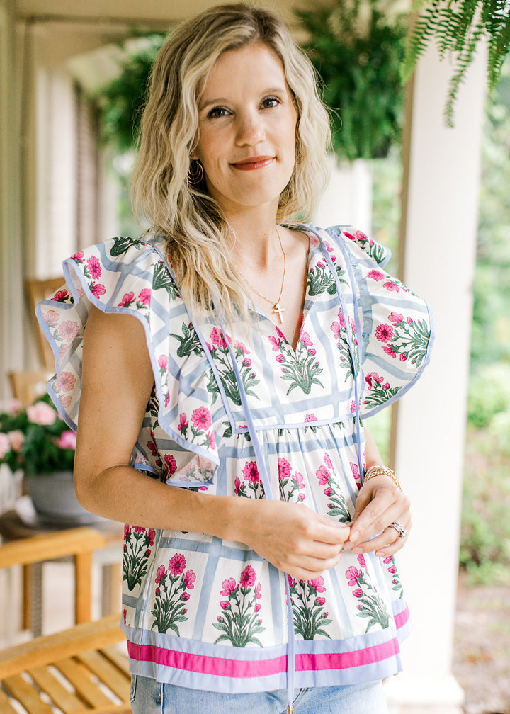 Model wearing an off white v-neck top with pale blue squares and pink flowers and ruffle sleeves.