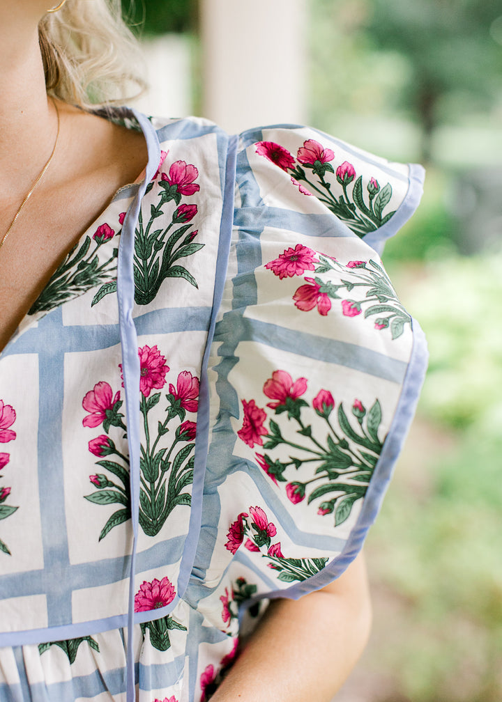Close up of ruffle sleeves and tie v-neck on an off white top with blue squares and pink flowers.