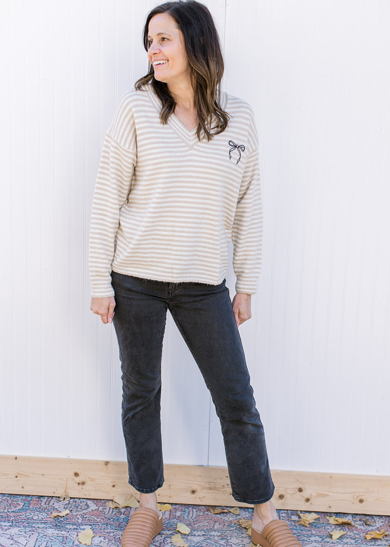 Model wearing black jeans, mules and a cream and tan sweater with a black bow and long sleeves.