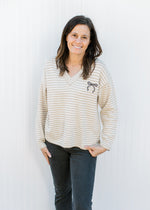 Model wearing a cream and tan v-neck sweater with a black graphic bow emblem and long sleeves.