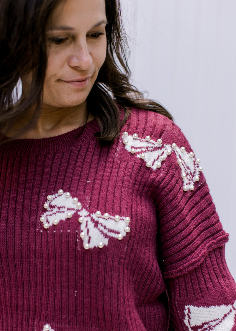 Close up of cream bows, embellished with pearl beads on a burgundy long sleeve sweater. 