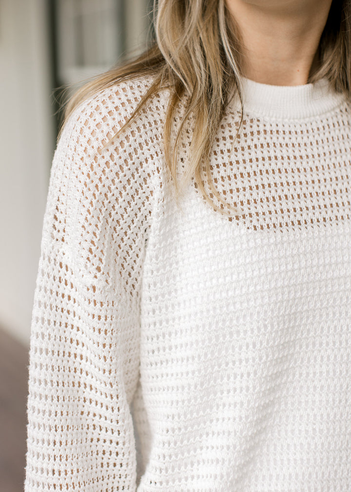 Model wearing a white, open-knit crochet top with a round neckline and long sleeves. 