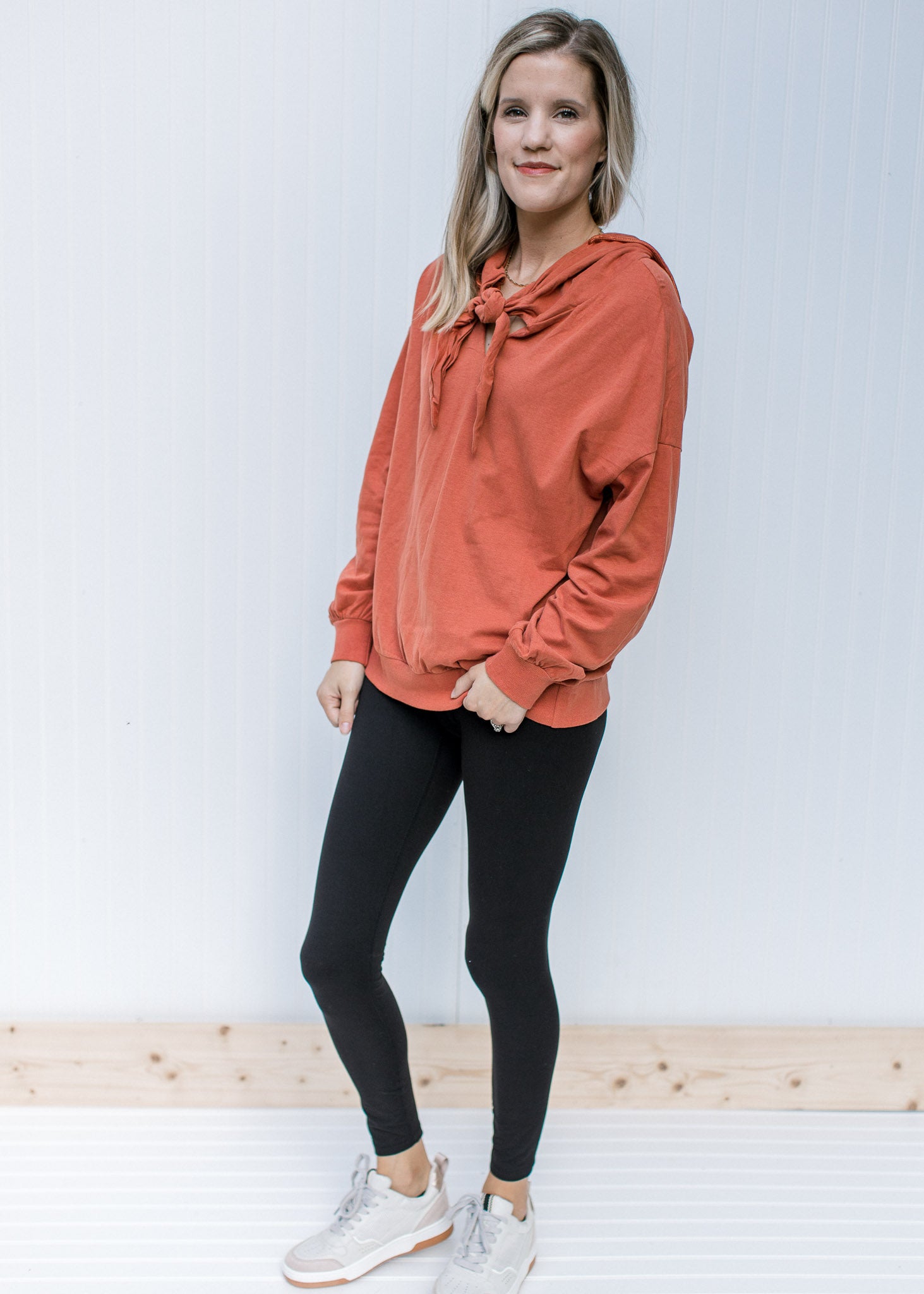Model wearing jeans with a brick long sleeve sweatshirt with a hood and v-neck with a tie. 