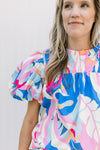 Model wearing bright floral top with a smocked yolk, ruffle neck and short puff sleeves. 