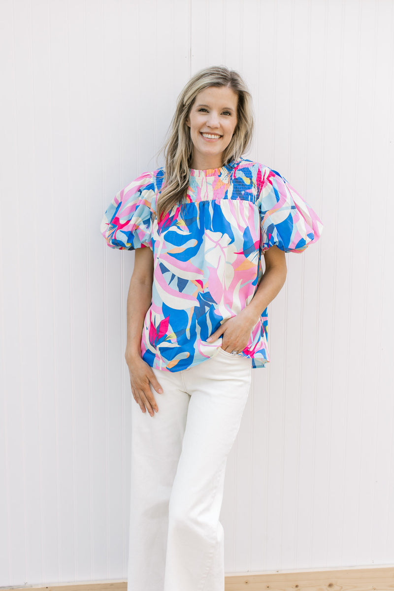 Model wearing white jeans with bright floral top with a smocked yolk, ruffle neck and short sleeves.