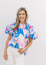 Model wearing bright floral top with a smocked yolk, ruffle neck and short sleeves.