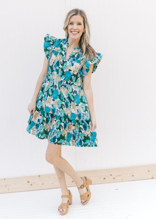 Model wearing heels with a tiered bright blue floral dress with ruffle cap sleeves and a v-neck.
